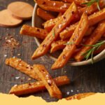 Pile of golden crispy oven baked sweet potato fries on a parchment-lined baking sheet with a small bowl of dipping sauce.
