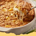 A golden-brown Easy Pineapple Cream Cheese Cobbler fresh from the oven, with a creamy, bubbly pineapple filling peeking through its cake crust. Served in a white baking dish.