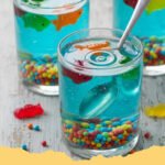 Edible Aquarium Jello Cups Recipe - Close-up of a vibrant Edible Aquarium Jello Cup with gummy fish swimming in blue jello, garnished with a sandy bottom and green seaweed.