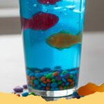 Edible Aquarium Jello Cups Recipe - Close-up of vibrant blue Edible Aquarium Jello Cups filled with candy fish and crushed graham cracker 'sand', ready to be served at a party.