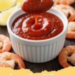 A vibrant red bowl of homemade cocktail sauce with fresh lemon slices and a few cooked shrimp on the side.