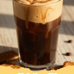 Close-up of a glass of iced coffee topped with a thick layer of cookie butter cold foam, garnished with crushed Biscoff cookies.
