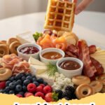 A beautifully arranged breakfast board overflowing with pancakes, waffles, fresh fruit, bacon, sausage, cheeses, pastries, and various spreads.