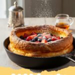 dutch baby recipe - A golden, puffed Dutch Baby pancake in a cast iron skillet, dusted with powdered sugar and garnished with fresh lemon wedges and blueberries.