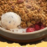 best rhubarb recipes - A golden brown rhubarb crumble in a ceramic baking dish, with a scoop of vanilla ice cream melting on top.