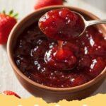 strawberry jam recipe - Close-up of vibrant red homemade strawberry jam in a glass jar, with fresh strawberries and a spoon beside it.