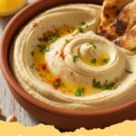 hummus recipe homemade - A bowl of creamy, homemade hummus drizzled with olive oil and garnished with paprika and parsley, served with pita bread.
