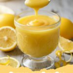 lemon curd recipe - A jar of vibrant yellow, silky smooth homemade lemon curd with a lemon slice and a spoon next to it.