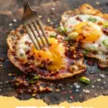 Chilli Oil Fried Eggs - Two perfectly cooked fried eggs with crispy edges and runny yolks, swimming in vibrant red chilli oil, garnished with green onions, served on a bed of white rice.