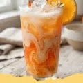A tall glass of Dirty Orange Creamsicle Soda with ice, an orange slice, and a straw, showing distinct creamy and orange layers.