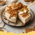 A slice of Greek Yogurt Biscoff High-Protein Cheesecake on a white plate, garnished with crumbled Biscoff cookies and a drizzle of Biscoff spread.