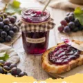grape jelly recipe - Several jars of vibrant purple homemade grape jelly on a wooden table, with fresh Concord grapes and leaves in the foreground.