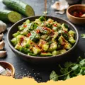 A vibrant bowl of Smashed Cucumber & Garlic Sesame Salad, featuring irregularly cut cucumbers, sesame seeds, and red chili flakes, glistening with a savory dressing.